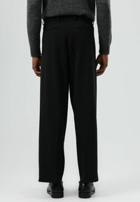 Zwarte, wijdvallende broek met een getailleerde pasvorm, gemaakt van een gladde stof. Voorzien van taillebandlussen en twee achterzakken. Gecombineerd met een grijze gebreide trui.