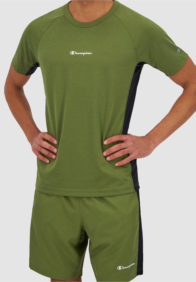 Homme portant un t-shirt de sport Champion vert olive et un short assorti avec des panneaux latéraux noirs, les mains reposant sur les hanches.