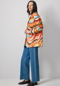 Colorful, patterned shirt in orange, blue, and brown waves; paired with wide-leg blue denim jeans. Model wears dark shoes.