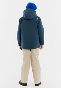 Blauwe waterdichte jas met een capuchon, elastische manchetten en een logo op de achterkant. Beigekleurige broek en zwarte laarzen maken de outdoor outfit compleet.
