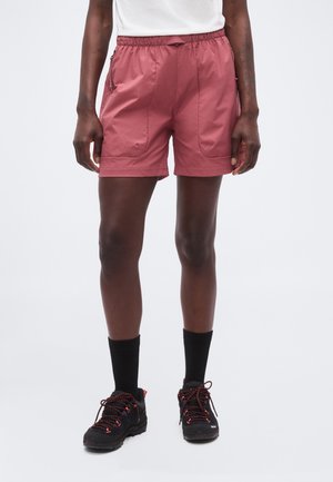Personne portant un short de sport rose, des chaussettes noires mi-mollet et des chaussures de randonnée noires avec des détails rouges, debout devant un fond uni.