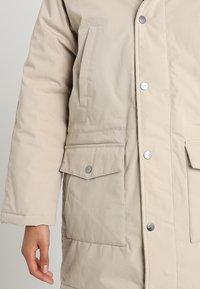 Abrigo acolchado beige con cierre de botón en la parte delantera, dos grandes bolsillos frontales y una textura suave. Presenta acentos de hardware metálico.