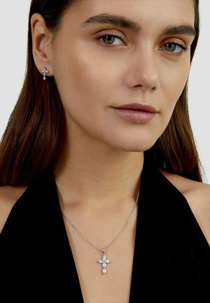 Frau mit langen braunen Haaren, die ein schwarzes Neckholder-Oberteil, eine Diamanten-Kreuz-Halskette und passende Diamanten-Kreuz-Ohrringe trägt.