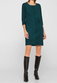 Femme portant une robe teal à mi-mollet avec des manches trois-quarts et des bottes en cuir noires jusqu'au genou, sur un fond clair.