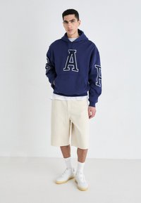 Felpa con cappuccio in felpa blu marina con grande stampa bianca a "A", abbinata a pantaloni corti beige fino al ginocchio e sneaker bianche, caratterizzata da una vestibilità rilassata e tasca a marsupio.