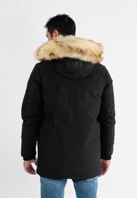 Parka noire avec une capuche bordée de fourrure, présentant un extérieur lisse, des manches longues et un dos légèrement plus long. Pas de fermetures ni de poches visibles.