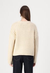 Cream-colored chunky knit sweater with a ribbed texture, featuring dropped shoulders and a rounded neckline, paired with dark denim jeans.