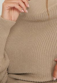 Beżowy sweter z dzianiny z wysokim, prążkowanym kołnierzem. Wykonany z teksturowanej, elastycznej tkaniny i dopasowanymi rękawami. Ręka spoczywająca na materiale.