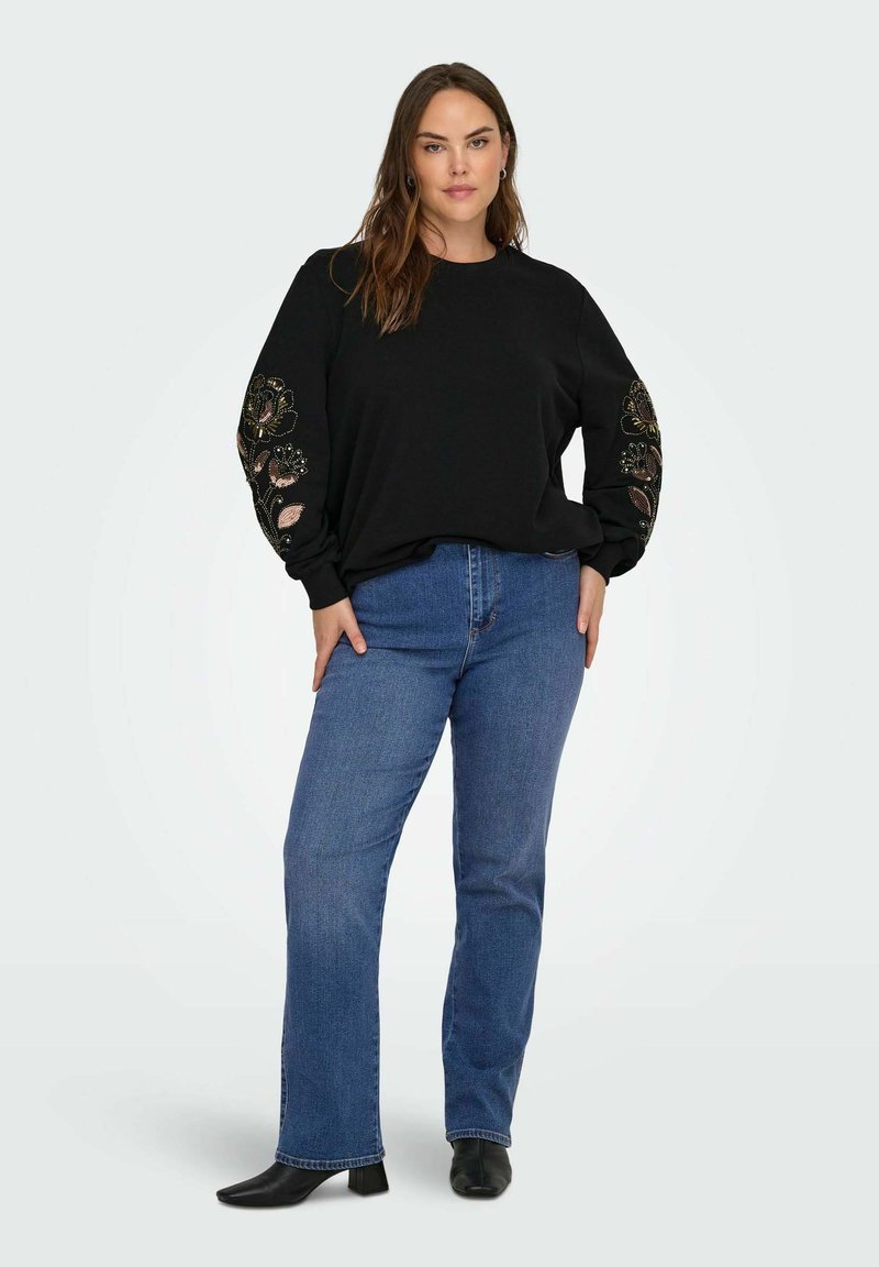 Crna majica sa cvjetnim vezom na rukavima, uparena s plavim trapericama ravnog kroja i crnim gležnjačama. Glatka tkanina i opušteni kroj.