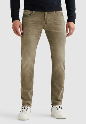 Hombre con jeans beige de corte ajustado, zapatillas blancas y un top negro de manga larga, de pie frente a un fondo liso.