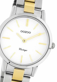 Montre en acier inoxydable avec un cadran blanc, des accents jaunes et un bracelet en métal argenté et doré. Elle présente des index horaires fins et un design vintage.
