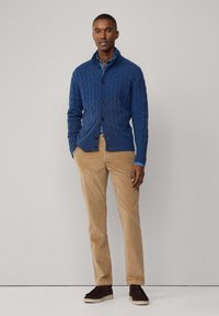 Blauwe kabelgebreide cardigan, dichtgeknoopt, gedragen over een lichtblauw overhemd met kraag. Tan veelkleurige corduroy broek en donkerbruine schoenen maken de outfit compleet.