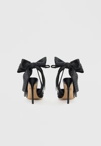 Pantofi cu toc înalt negri, cu finisaj lucios, având un design cu degetul la vedere și funda largă decorativă împodobită cu strasuri.