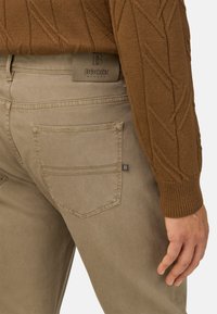 Beżowe spodnie z tylną kieszenią, z subtelnym przeszyciem. Tkanina ma gładką fakturę, w zestawieniu z brązowym swetrem dzianinowym.