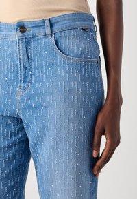 Nærbilde av en person som har på seg lyseblå denim jeans med sølvfarget paljett detaljer og en beige topp, hånden hviler ved siden.