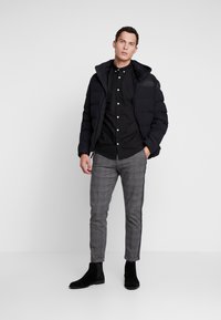 Zwarte pufferjack met capuchon, langemouw zwarte overhemd met knopen, grijs geruite broek, en zwarte enkelboots, staand tegen een neutrale achtergrond.