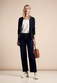 Blazer marine, haut blanc, jeans larges bleu foncé, et baskets blanches. Sac à main marron avec une courte anse. Design simple et épuré.