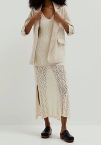 Blazer beige sopra un vestito maxi in pizzo lavorato a maglia crema, con motivo a diamante e spacco laterale; abbinato a zoccoli neri con suola in legno.