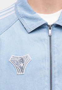 Ανοιχτό μπλε μπουφάν με φερμουάρ και γιακά, άσπρη μπλούζα από μέσα και μια ασημένια καρφίτσα σε σχήμα εσωρούχου καρφωμένη στην αριστερή πλευρά του στήθους.