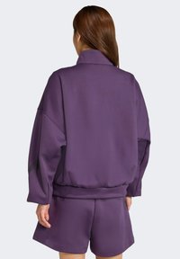 Prevelký fialový mikin s vysokým golierom a dlhými rukávmi, vyrobený z textúrovanej látky a s voľným strihom, skombinovaný s ladícími šortkami.