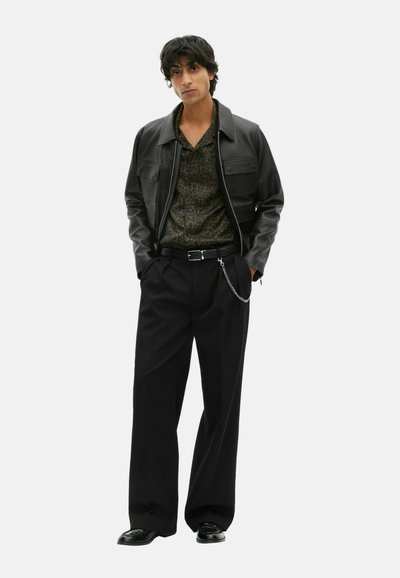 Black leather jacket over a patterned dark shirt, paired with loose black trousers and a silver chain accent on the belt.
