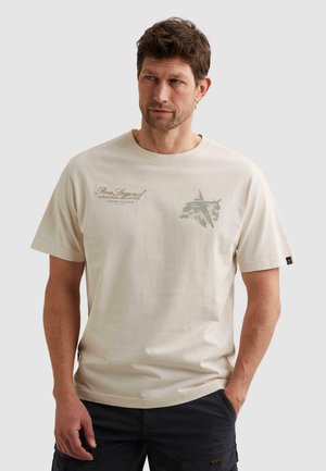 Man wearing beige short-sleeve T-shirt with "Pme Legend" text and cargo plane graphic, paired with black cargo pants, standing with one hand in pocket.