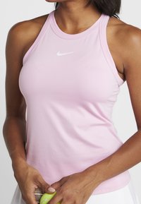 Różowy sportowy top na ramiączkach z teksturowanej tkaniny, zapinany na plecach, z białym logo Nike na piersi, trzymany przez osobę z piłką tenisową.