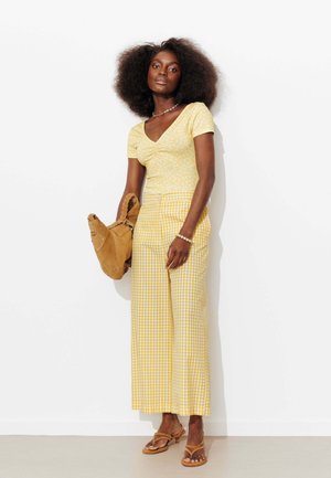 Femme aux grands cheveux bouclés porte un haut jaune à fleurs, un pantalon large à carreaux jaune, des sandales beige et tient un sac à main beige, debout contre un mur blanc.