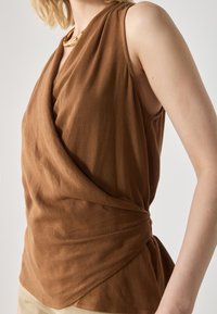 Braunes ärmelloses Oberteil mit drapiertem Design, das einen Wickeleffekt und strukturierte Stoffe aufweist. Abgerundet durch eine seitliche Drehung und einen lässigen Schnitt.