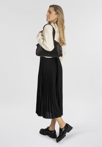 Zwarte geplooide midi-jurk, crèmekleurige sweater en glanzende zwarte platform schoenen; model houdt een zwarte leren schoudertas vast met trekkoorddetails.