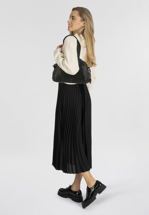 Zwarte geplooide midi-jurk, crèmekleurige sweater en glanzende zwarte platform schoenen; model houdt een zwarte leren schoudertas vast met trekkoorddetails.