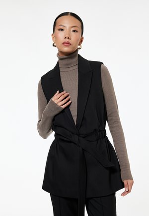 Vrouw draagt een taupekleurige coltrui en een zwart mouwloos gilet met ceintuur, staand tegen een witte achtergrond.