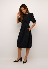 Robe noire, longueur genou, avec des manches bouffantes courtes, des poches latérales et une silhouette droite. Tissu lisse avec un léger éclat et des fermetures en or.