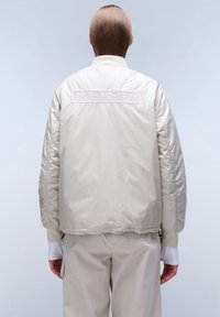 Beige bomberjack met een glanzende textuur, geribbelde manchetten en een geborduurd "NAPAPIJRI" logo op de achterkant. Smooth stof, relaxte pasvorm.