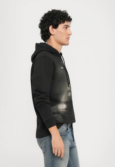 Zwarte hoodie met een grafische print van een auto op de borst, voorzien van een trekkoordkap en geribbelde manchetten. Gekozen in combinatie met lichtblauwe jeans.