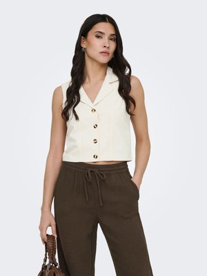 Donna con gilet crema senza maniche con bottoni e pantaloni marroni larghi con coulisse, che tiene in una mano una borsa marrone intrecciata.