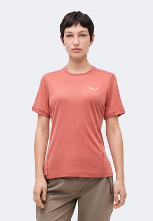 Woman with short dark hair wearing a salmon-colored short-sleeve t-shirt and beige pants, standing against a plain light background.