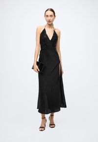 Black halter-neck dress with sequin detailing on the bodice, thigh-high slit, and flowing skirt; paired with a black clutch and strappy heels.