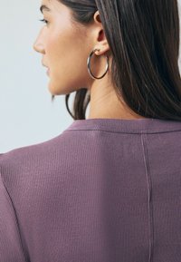 Purple ribbed fabric top with a low collar, showing a close-up of the back. Features a smooth texture and circular silver hoop earring.