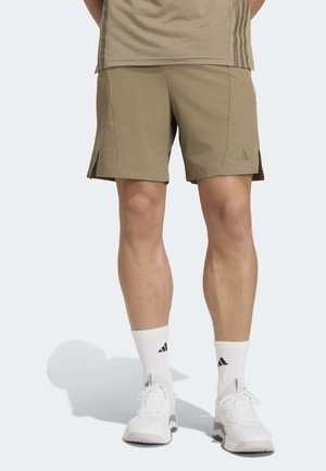 Mann trägt beige Sportshorts, weiße Sportsocken und weiße Turnschuhe und steht auf hellem Hintergrund.