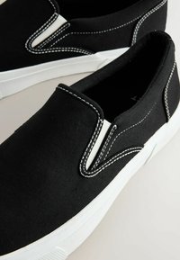 Schwarze Leinenschlupfschuhe mit weißen Nähten, elastischen seitlichen Einsätzen und einer strukturierten Gummisohle. Traditionelles Design mit minimalistischem Look.
