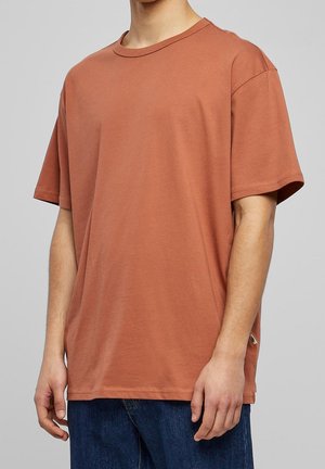 Mand iført løs pasform kortærmet rustorange t-shirt og mørkeblå jeans, stående mod en ensartet lysegrå baggrund.