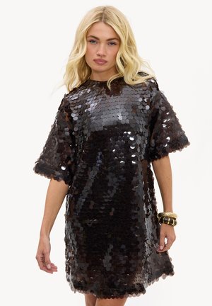 Femme blonde portant une robe noire ample couverte de grands sequins brillants avec des bracelets dorés et noirs à son poignet droit.