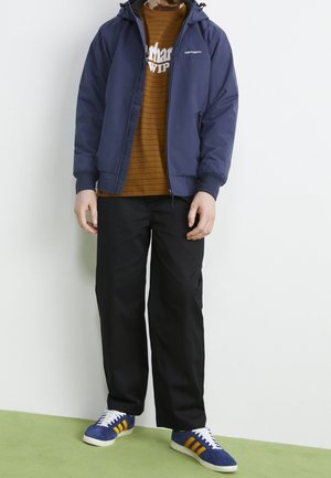 Osoba ubrana w granatową kurtkę Carhartt na brązowej koszuli w paski, czarne spodnie i niebieskie trampki z żółtymi paskami, stojąca na zielonej podłodze.