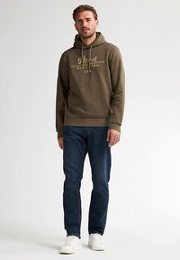 Olive zielona bluza z kapturem z grafiką z przodu w kolorze złotym, kapturem ze sznurkiem, ściągaczami na mankietach i u dołu. Połączona z ciemnymi dżinsami i białymi trampkami.