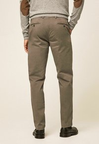 Muž oblečený v hnědých slim-fit kalhotách, šedém svetru s hnědými loketními záplaty a černých společenských botách, stojící zády k béžovému pozadí.