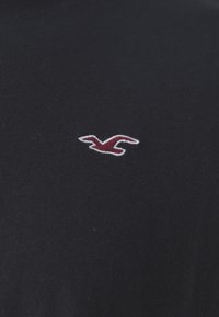 Melnā kokvilnas t-krekls ar mazu sarkanu iešūto logo kreisajā krūtī. Audums šķiet mīksts ar gludu tekstūru.