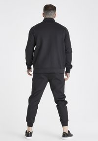 Felpa nera con collo alto, pantaloni jogger neri aderenti e scarpe da ginnastica nere. Tessuto liscio, vestibilità comoda, design minimalista.