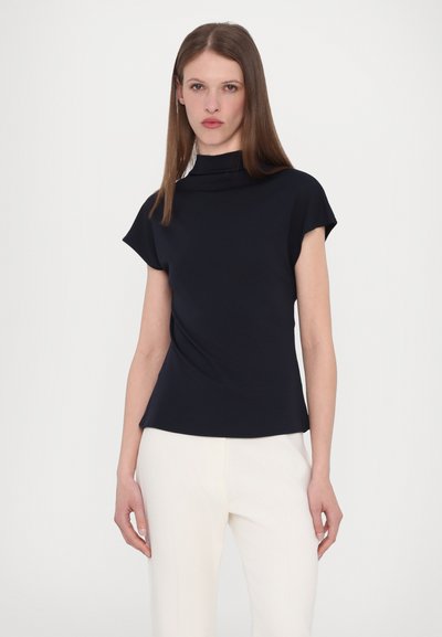 Marineblauwe blouse met korte mouwen en een hoge halslijn, gladde textuur en een ontspannen pasvorm. Gedragen met lichte broek. Minimalistisch ontwerp.