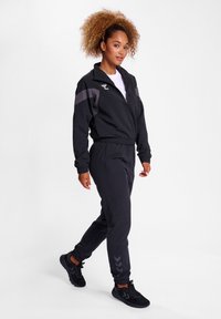 Svart träningsdräkt i mjukt tyg, med en zip-up jacka med hög krage och matchande byxor med muddar. Förstärkt med grå ränder.